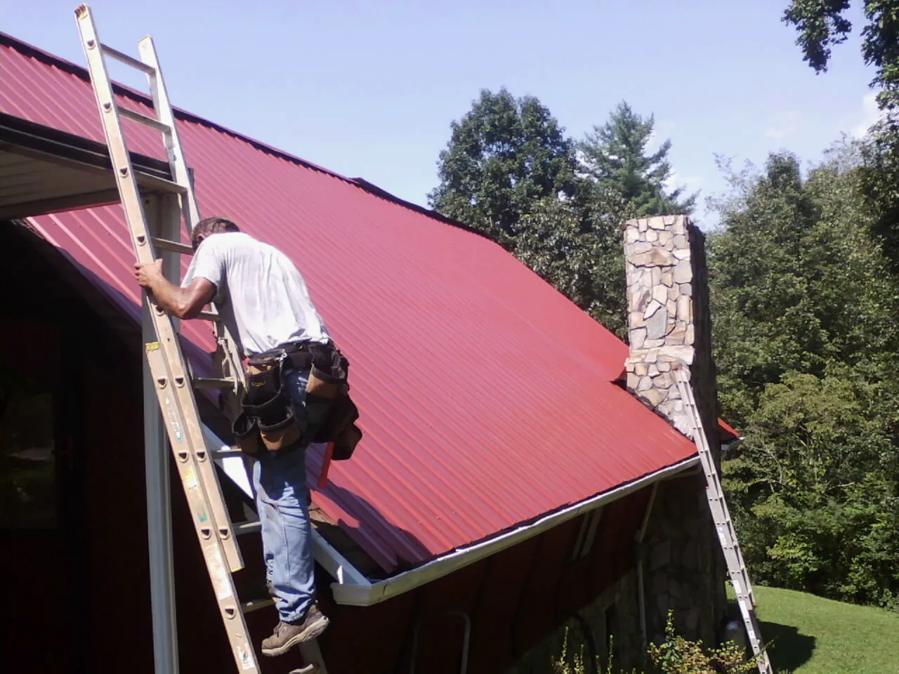 Roofer on a ladder beside a red metal roof and stone chimney — Soffit & Fascia Repair by Roofing Brothers in Lurgan